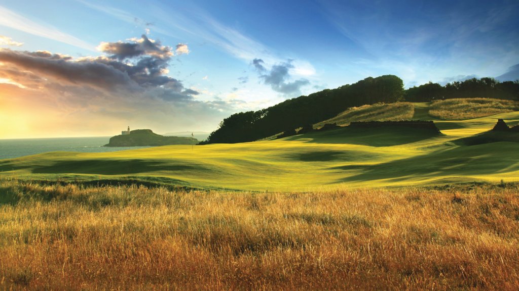 A scenic view of a golf course, likely a links course, with rolling green fairways and tall, golden fescue grasses in the foreground. In the distance, a body of water (ocean) meets the sky, with a prominent lighthouse on a rocky outcrop. The sky is partly cloudy with a warm, golden light suggesting either sunrise or sunset.